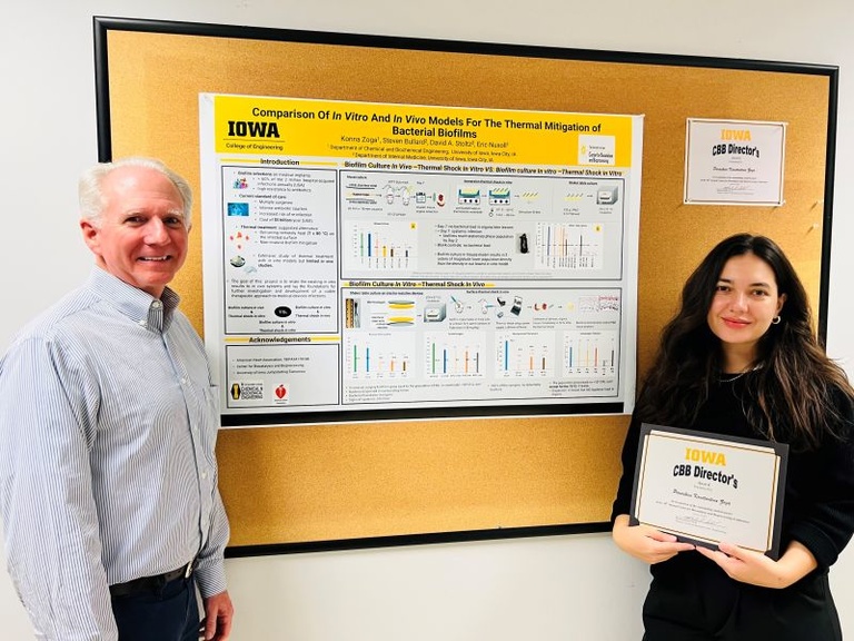 Konna Zoga stands to the right of her research poster. She is wearing a black outfit and holding an award for her research. A man in a white and blue plaid shirt stands to the left of her poster.