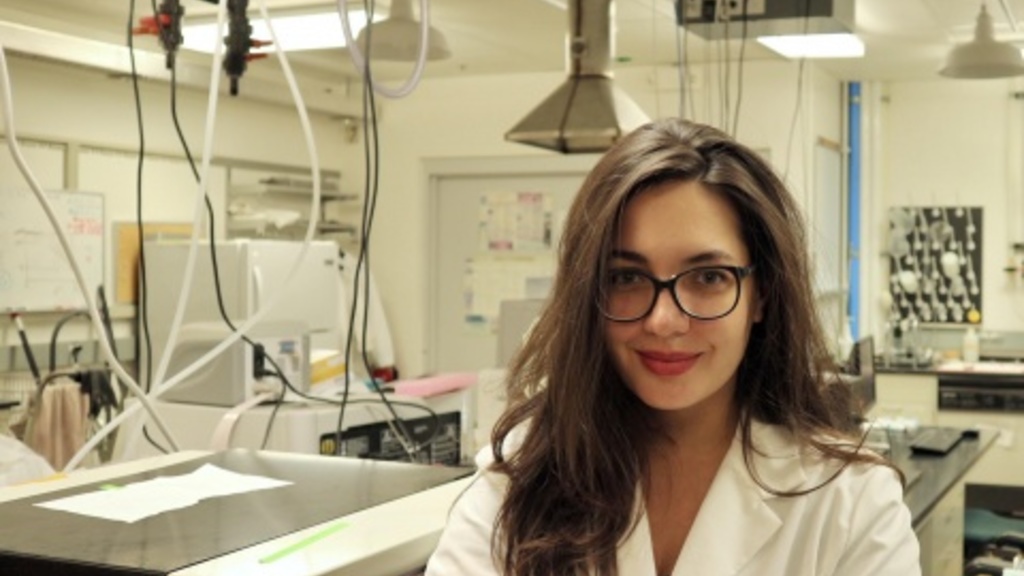 Konna Zoga stands with her arms folded and a smile on her face in front of her lab bench. She wears a white coat and black glasses.