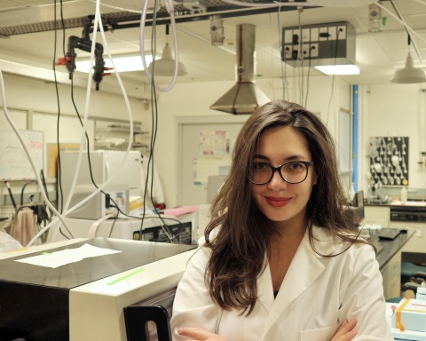 Konna Zoga stands with her arms folded and a smile on her face in front of her lab bench. She wears a white coat and black glasses.