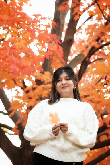 A portrait of Nuzhat Nuari Khan Rivu standing in front of orange trees. Rivu wear a white sweater and hold a light orange leaf in her hand.