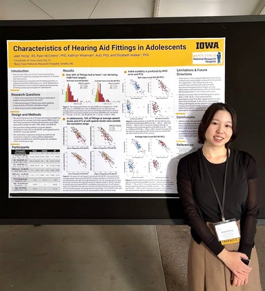 Jean Hong stands to the right of her poster at a presentation. She wears a black top and khaki pants as well as a white nametag. Her poster is Iowa branded and titled "Characteristics of Hearing Aid Fittings in Adolescents"