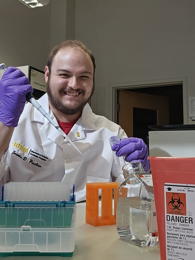 Travis sits at a lab bench in his white coat. He wears purple gloves and holds pieces of lab equipment up smiling at the camera.