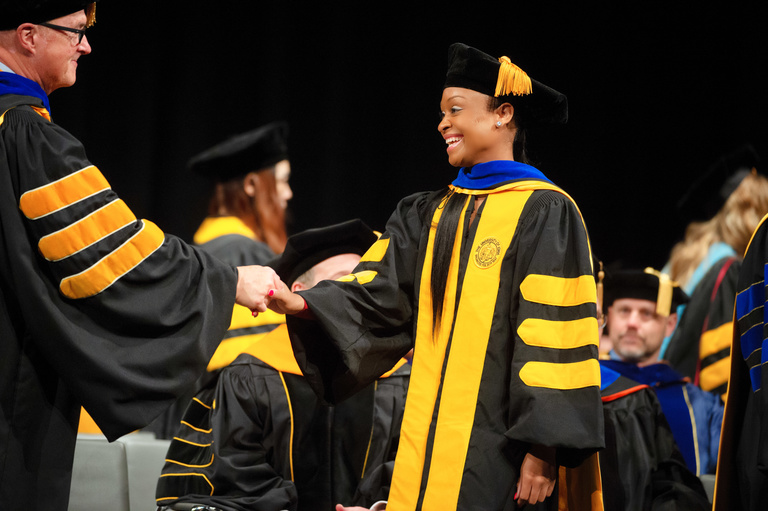Woman in doctoral regalia shaking a man's hand as she walks across commencement stage