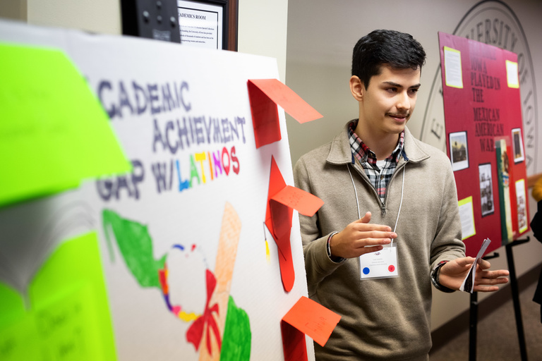 Picture of male student standing to the right of his poster titled "Academic Achievement Gap with Latinos"