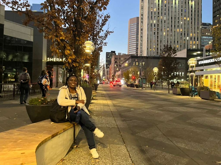 Cecilia sits on a bench on a street. She wears a white top with dark bottoms and white shoes. Her legs are crossed and she smiles toward the camera.