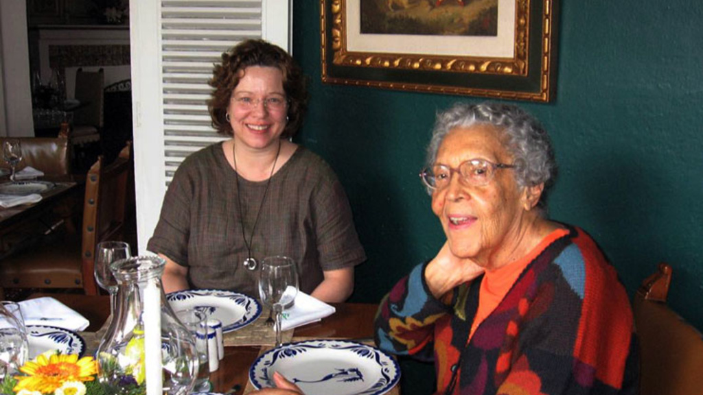 Elizabeth Catlett and another woman sit at a dining table and smile at the camera